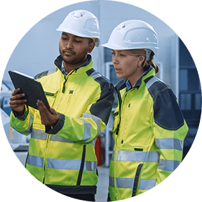 Two workers (male and female) in safety vests and hard hats looking at laptop on job site